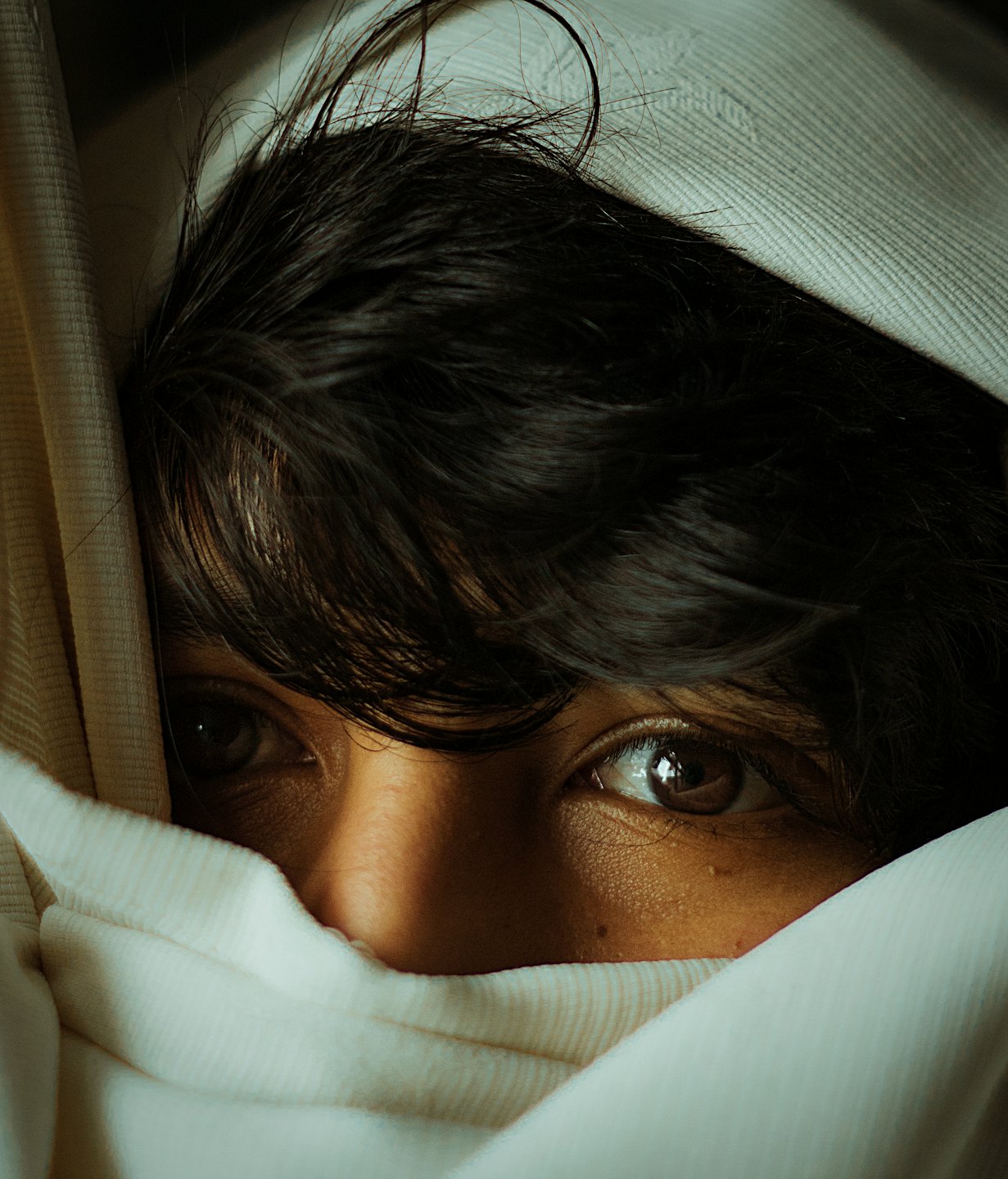 Person with dark hair gazes intently from behind cream fabric.